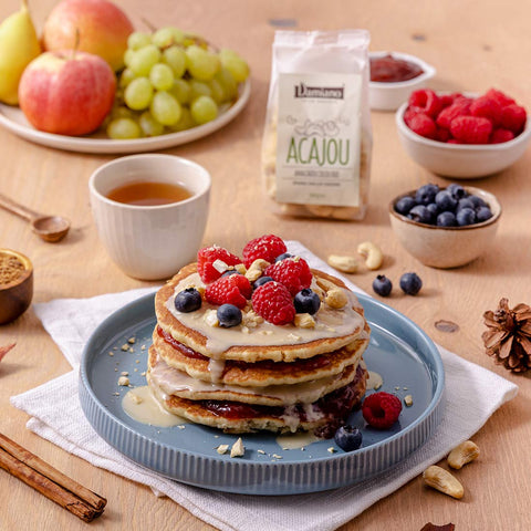Una colazione golosa e fresca con quattro pancakes impilati con marmellata di lamponi, crema di anacardi bianca e pezzetti di anacardi croccanti. Sul tavolo ci sono anche un tè caldo e tanta frutta.